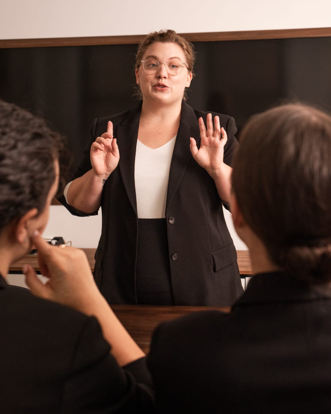 woman wearing black blazer and white top, standing in front of a chalkboard lecturing two people sitting in front of her