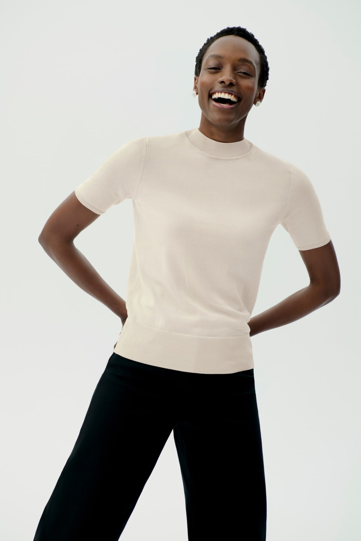 woman in front of light grey background, hands behind her back and smiling, wearing white short-sleeve top and black jeans