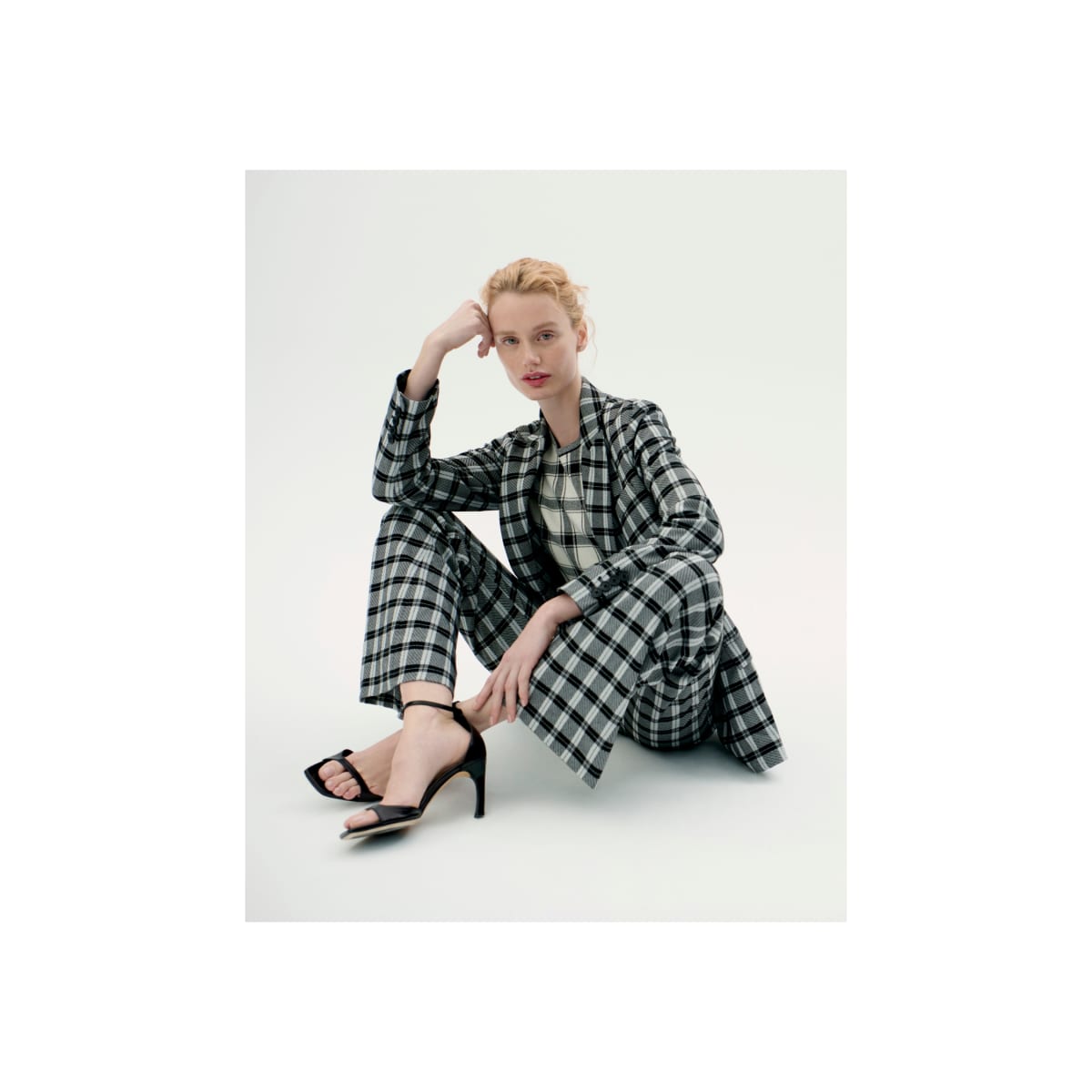 woman sitting with one elbow on one knee in front of light grey background, wearing black/white check plaid suit