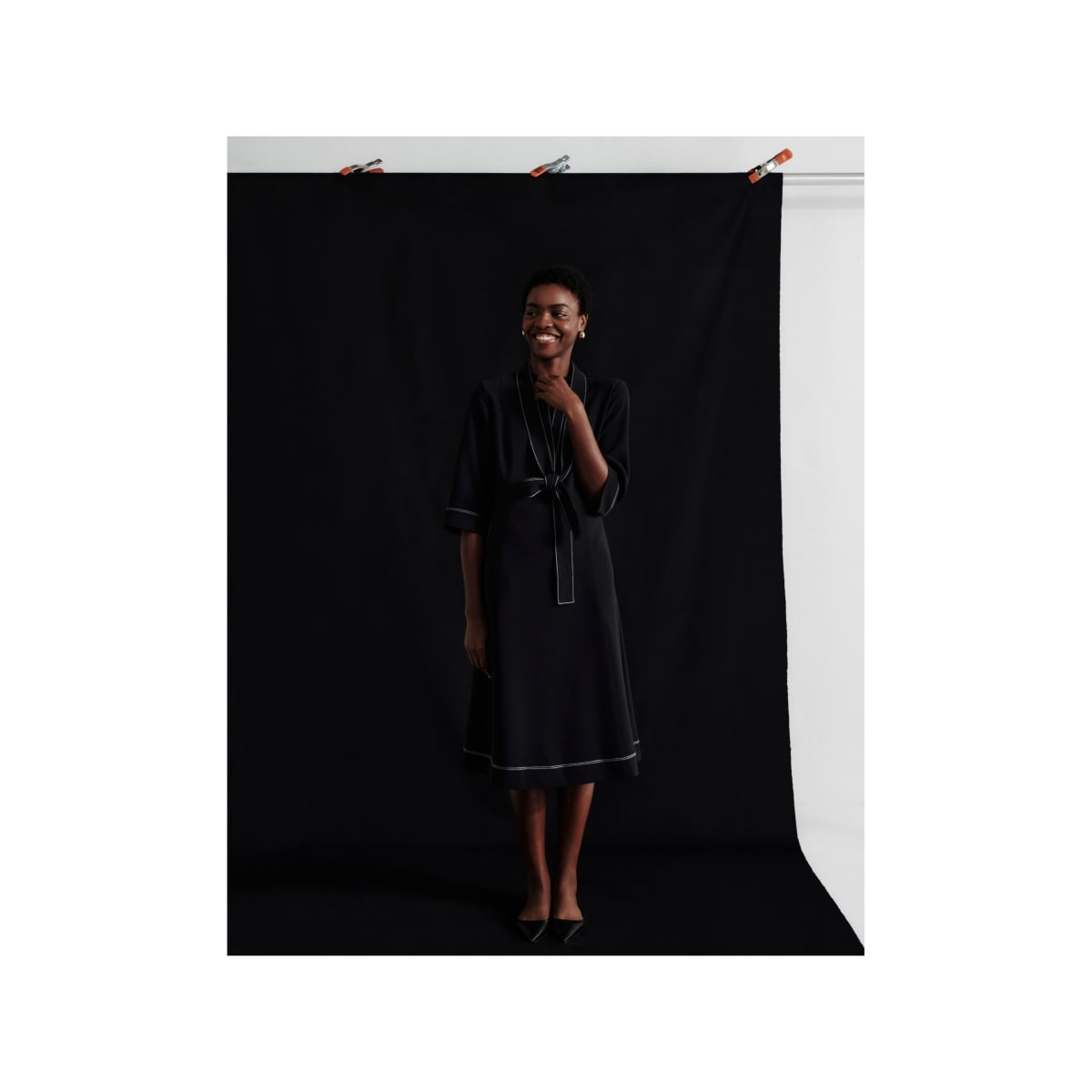 woman standing in front of black backdrop, wearing black midi-length mid-sleeve Lauleh dress with white contrast stitching