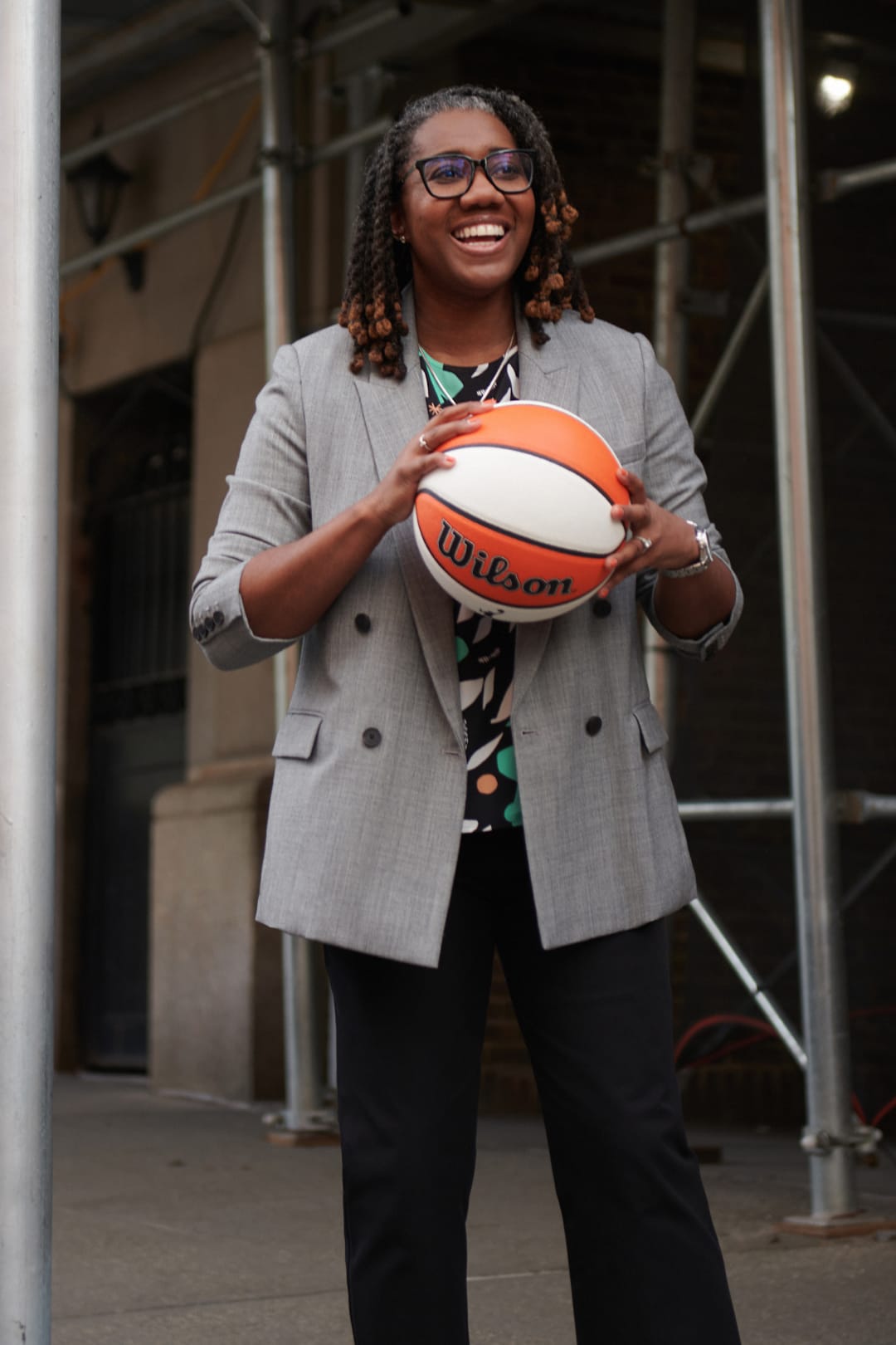 Keia Clarke smiling and holding a basketball and wearing M.M.LaFleur gray double-breasted blazer and black pants