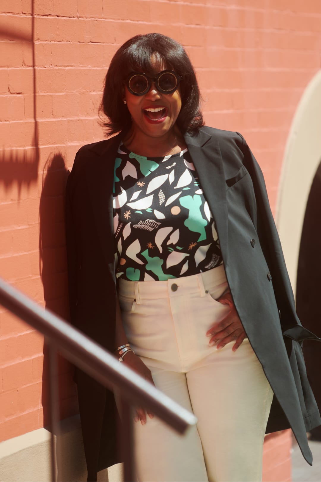 Tiffany Joy Murchison, wearing sunglasses and smiling to the camera, wearing black M.M.LaFleur coat over her shoulders, Didion top in black/white/seafoam green icon print, and white jeans
