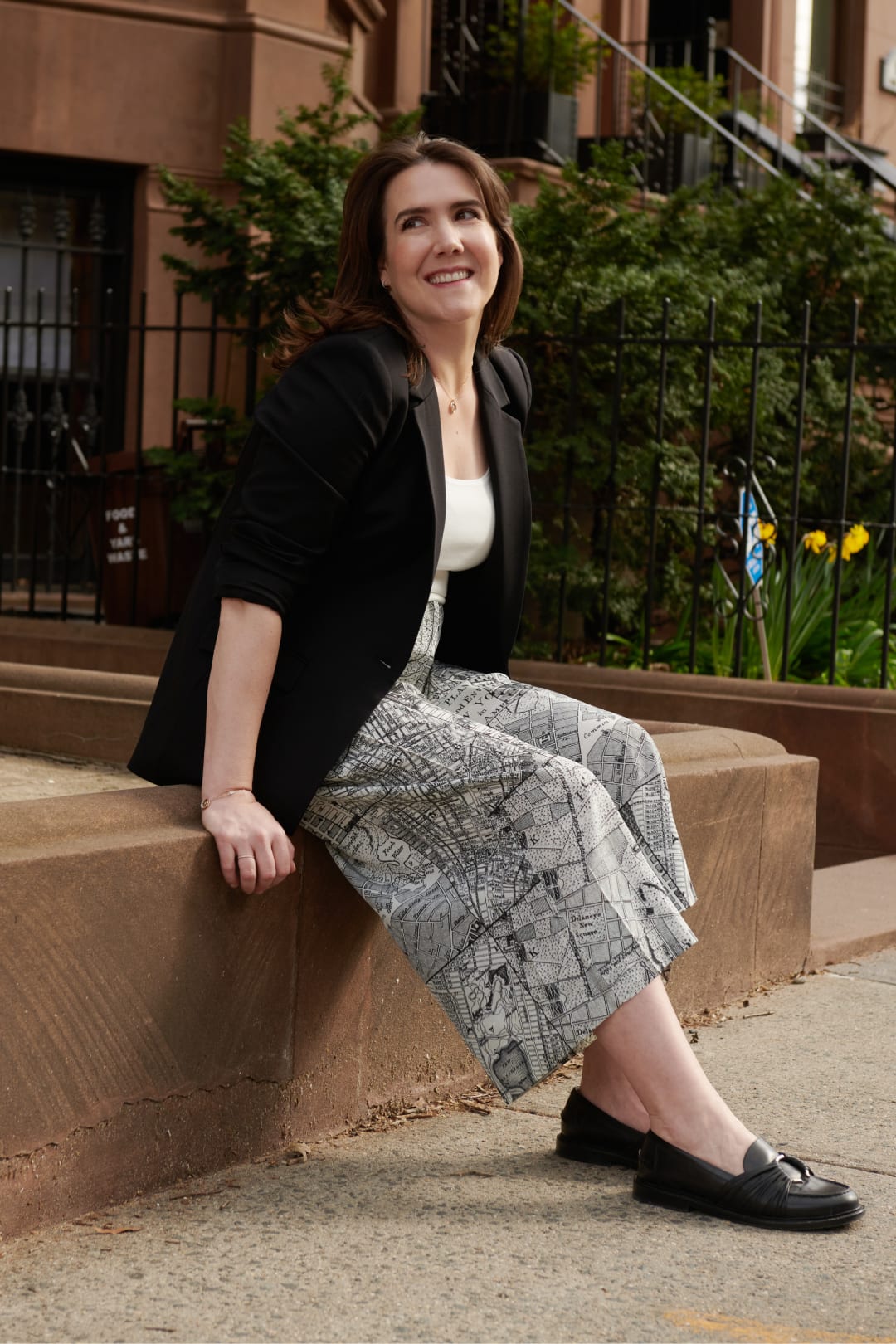 Maribeth Gainard sitting on a bench, and looking off-camera, wearing black M.M.LaFleur jacket, white top, culottes with the borough-map print, and black loafers
