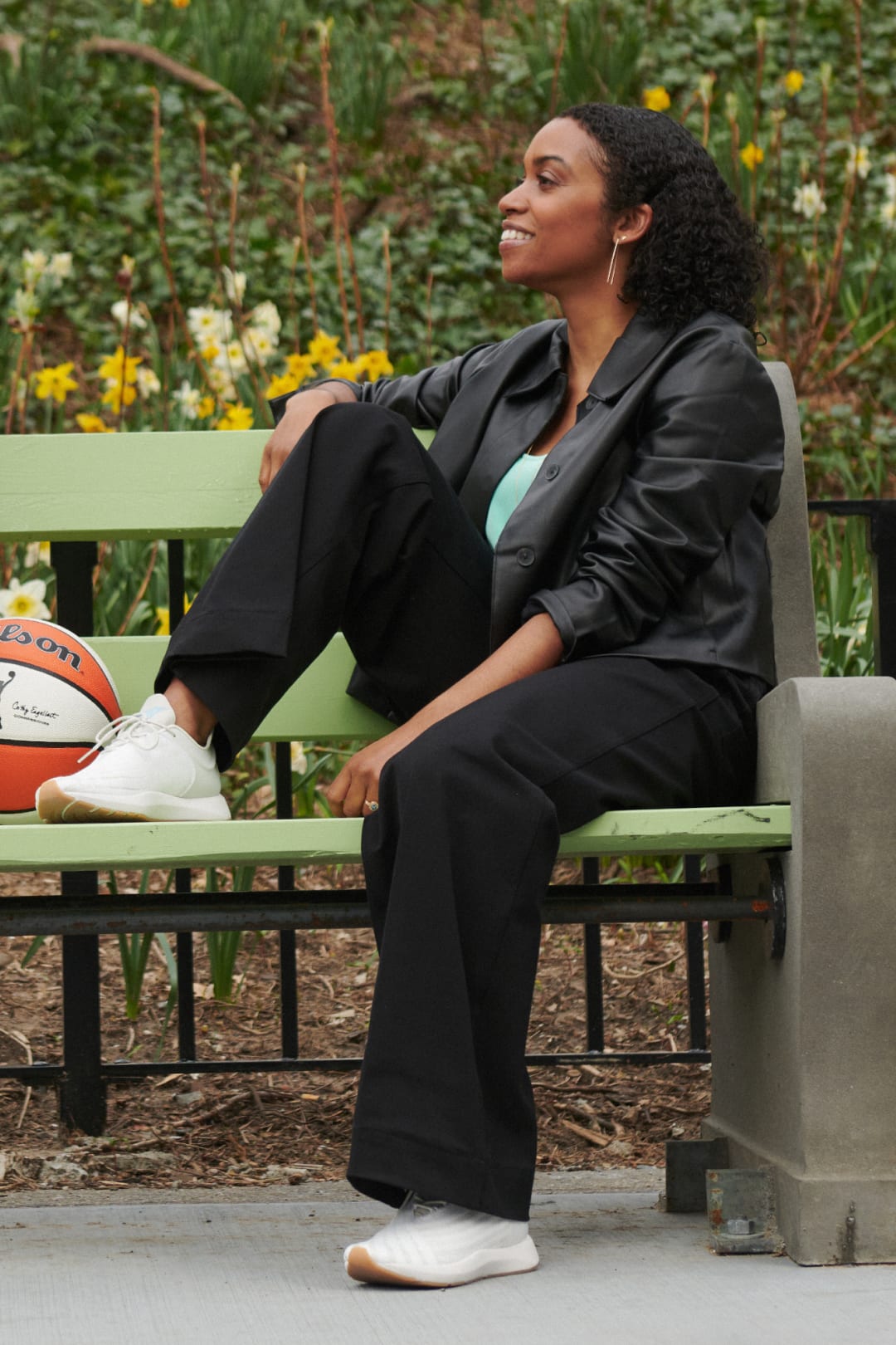 Keia Cole, sitting on a green bench outside with one leg on the bench, wearing black M.M.LaFleur vegan leather jacket, seafoam green T-shirt, black pants, and sneakers with a basketball next to her