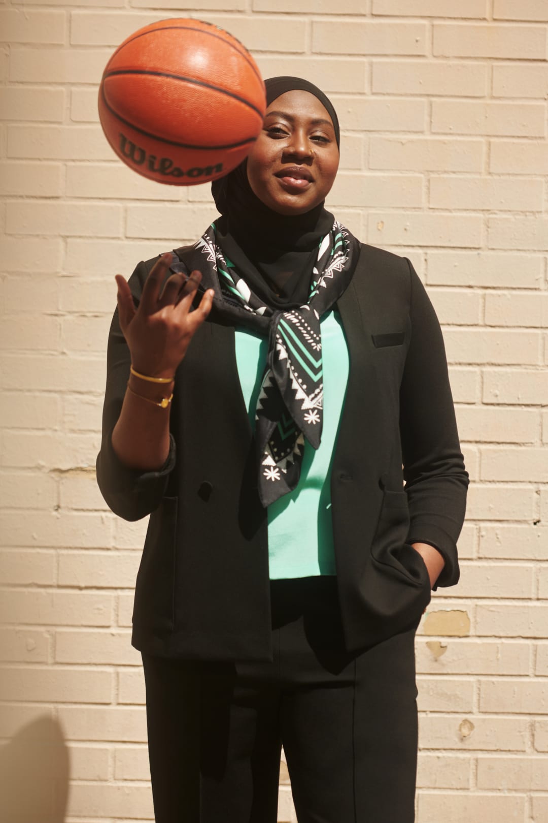 Batouly Camara M.S., wearing black hijab, black M.M.LaFleur blazer, seafoam green T-shirt, black pants, and black/white/seafoam icon print scarf tied around her neck, with one hand in pocket and the other throwing a basketball in the air