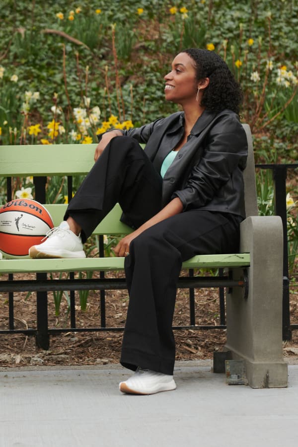 woman wearing M.M.LaFleur black vegan leather Nicky jacket with seafoam-colored Vicky tank underneath, sitting on a green bench outdoors with one foot propped up on bench