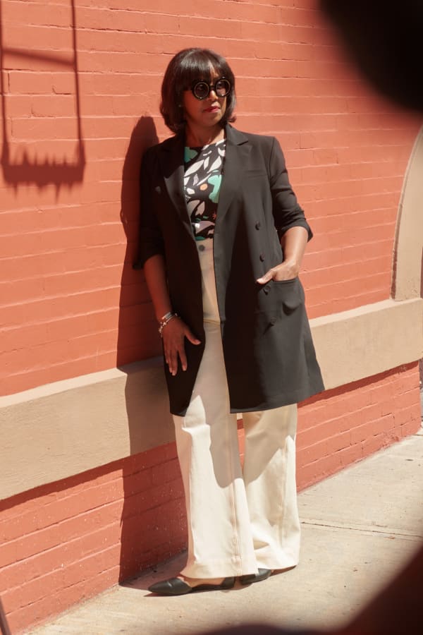 woman leaning against a brick wall outside, smiling and wearing sunglasses, M.M.LaFleur black jacket over her shoulders, Didion top in icon print, and white wide-leg Milo jeans