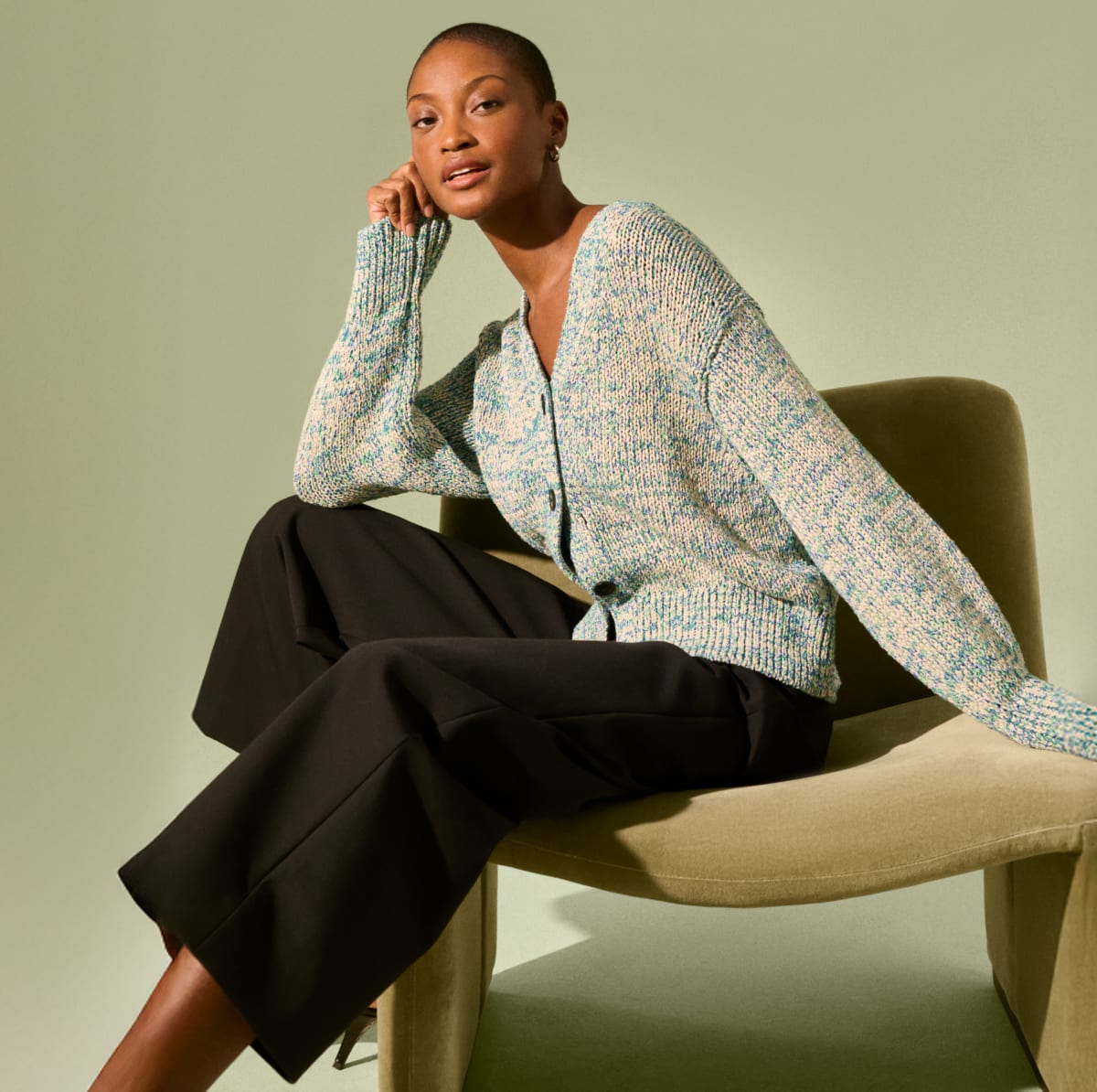 Image of a woman wearing the Dakota cardigan sitting in a green suede chair 