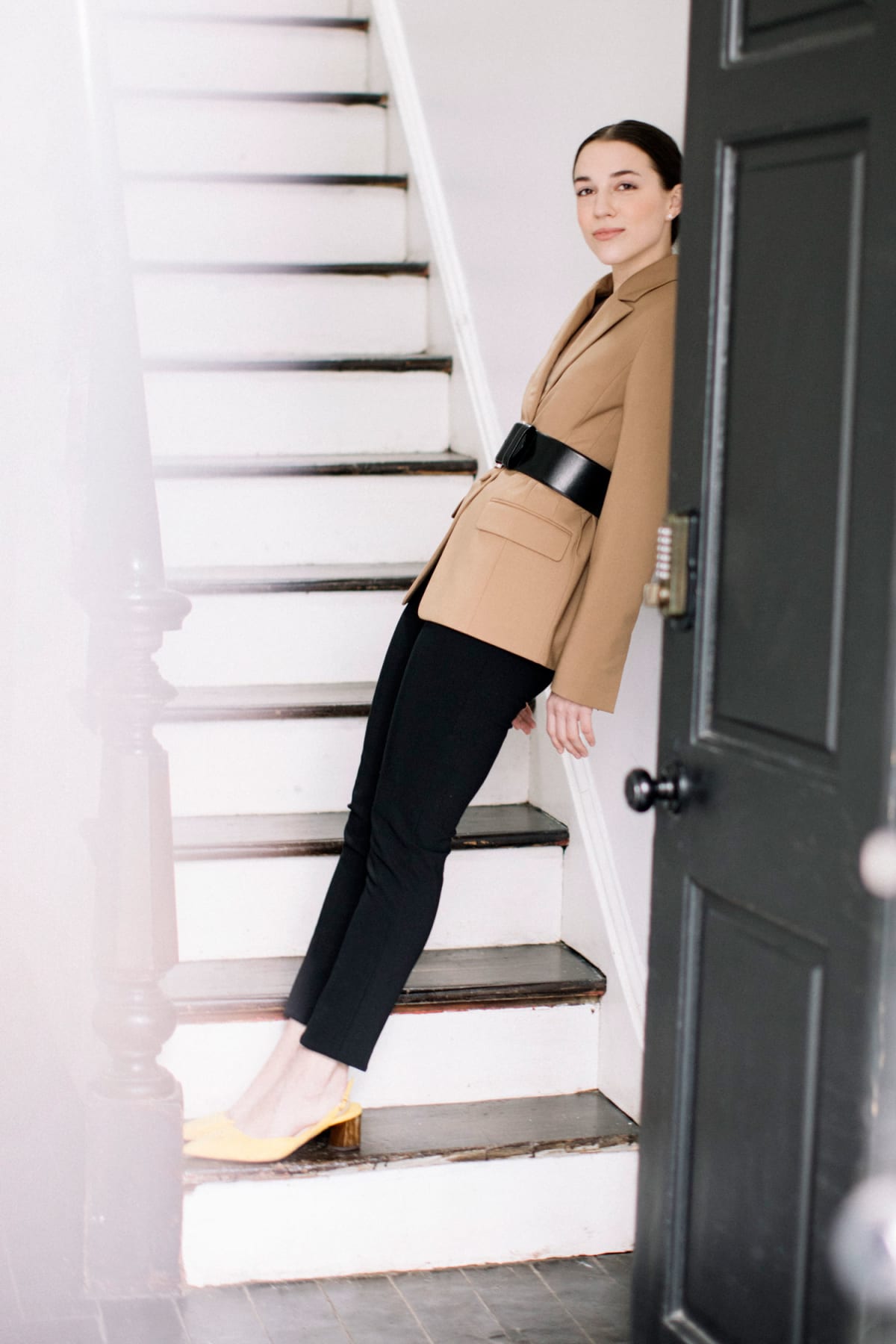 Woman on staircase wearing belted camel jacket over black skinny jeans