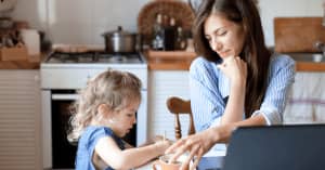 A mother and her toddler daughter are together at the kitchen table. The mother sits and smiles at the little girl