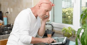 A middle-aged man stands in the kitchen and looks at his iPad and talks on the phone