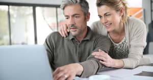 A man looks at a computer while a woman hugs him from behind and looks at the computer over his shoulder