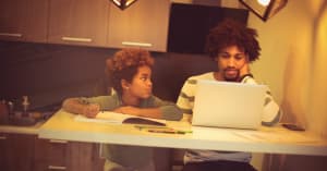 A little girl sits at a counter in the kitchen looking concerned while her dad looks at a computer and seems stressed out