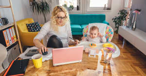 A woman looks at her home finances and investments on her computer while her baby sits in a highchair next to her