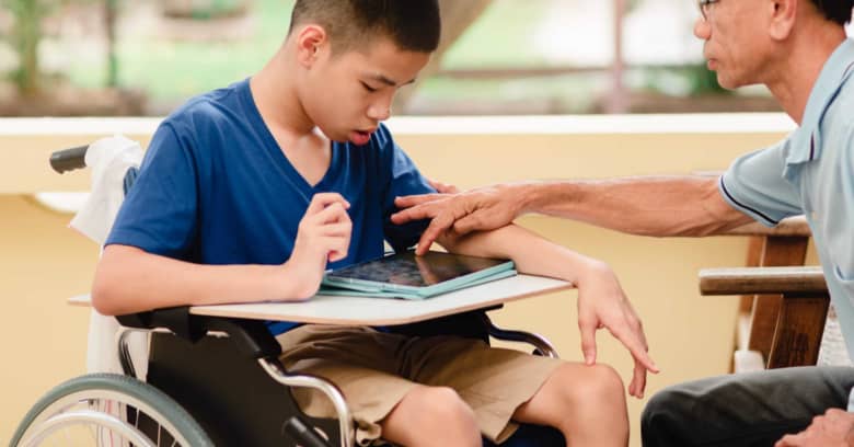 A student using a wheelchair looks at a computer tablet with his caretaker.