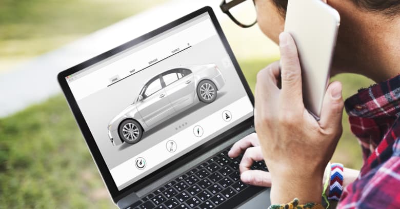 Looking over a man's shoulder, you can see he is researching cars for sale on his computer and he is also talking on the phone to ask about a car he sees