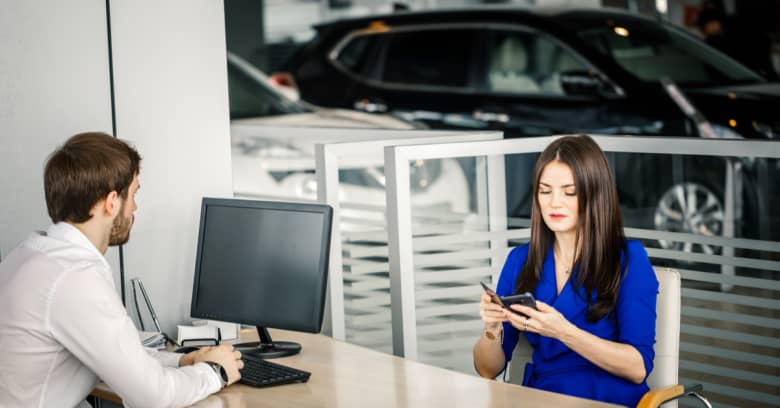 a woman sits at a desk at a car dealership with a salesman as she negotiates the price of a car she's interested in buying