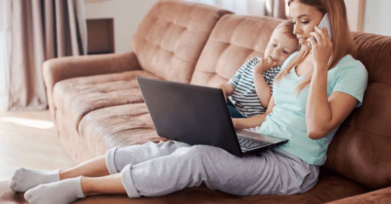 A woman sits on her couch, with her toddler child next to her, while speaking on the phone to her insurance agent