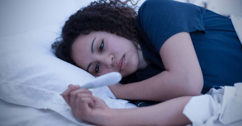 A woman lies on her side in bed looking at a thermometer, and she looks like she does not feel well