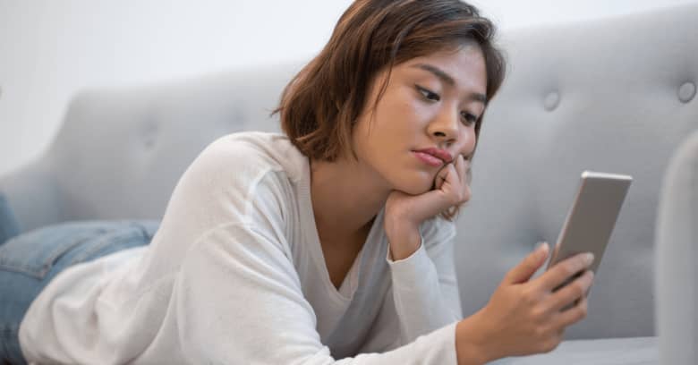 a woman with a bored expression lays on the couch and looks at her phone.