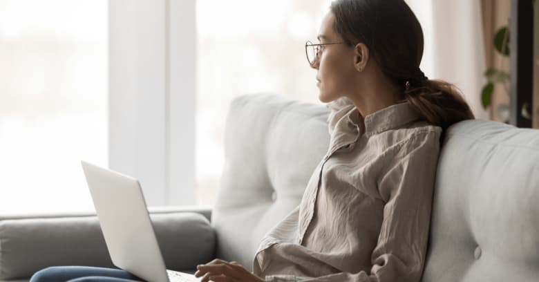 a woman sits on the couch with a laptop computer on her lap. She is gazing distractedly out the window.