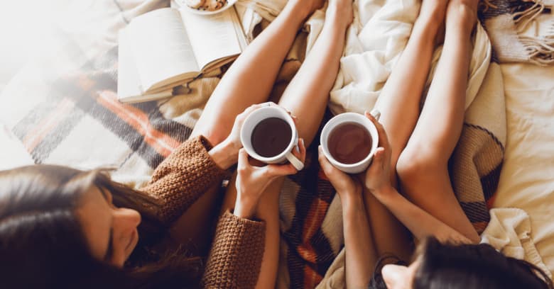 Two sisters sit on the bed with a book nearby as they enjoy a cup of tea together