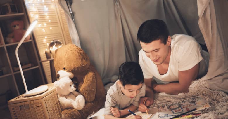 A father and his son draw pictures together as they lay on their stomachs in a living room fort they built