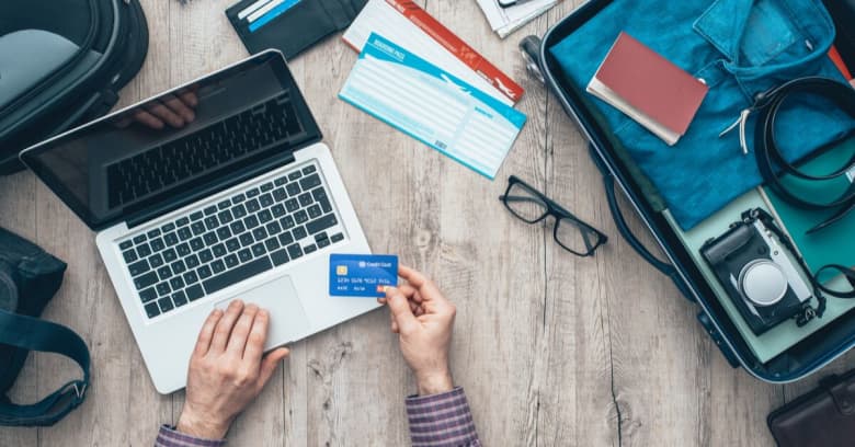 A man's hands holding a credit card in one hand while using the trackpad on his laptop with other preparing to cancel a trip that he is already packed for.