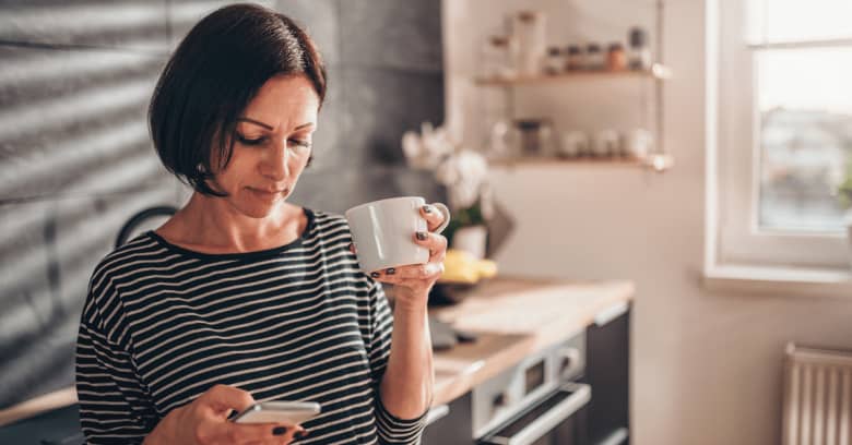 A woman stands in a kitchen with her phone and a cup of coffee.