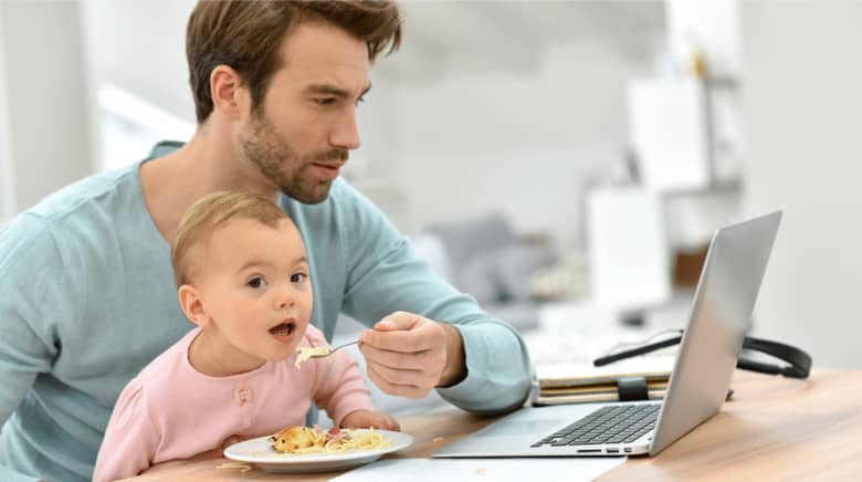 A man is working on his laptop and trying to feed his baby at the same time