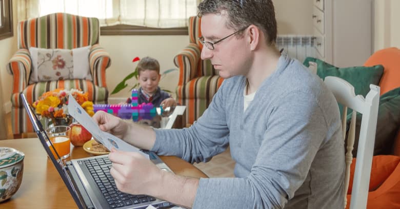 a man sits at a table while working on his computer while his son plays with colorful blocks in the background