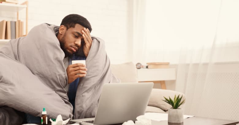A man sits at home sick with a blanket wrapped around him and a computer in front of him