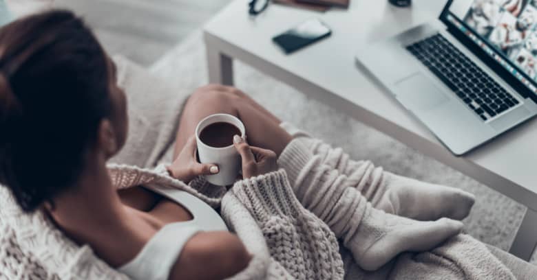 a woman has a cup of coffee while sitting on the couch looking at her computer on the coffee table. She is shown from above