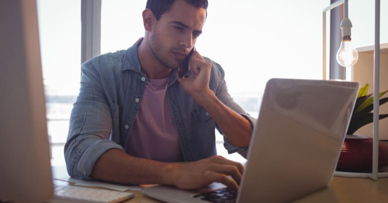 a man looks concerned as he talks on the phone and looks at his computer