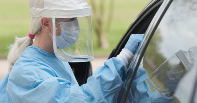 a woman in protective gear gives an occupant of a car a coronavirus test in a drive-through testing location