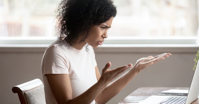 a woman looks at her computer with an incredulous look on her face using frustrated hand gestures