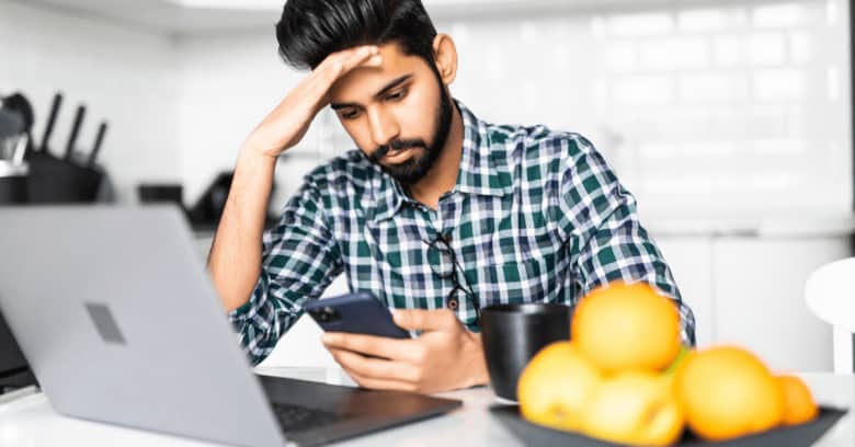 A man sits in his kitchen looking at his computer with a tired expression on his face