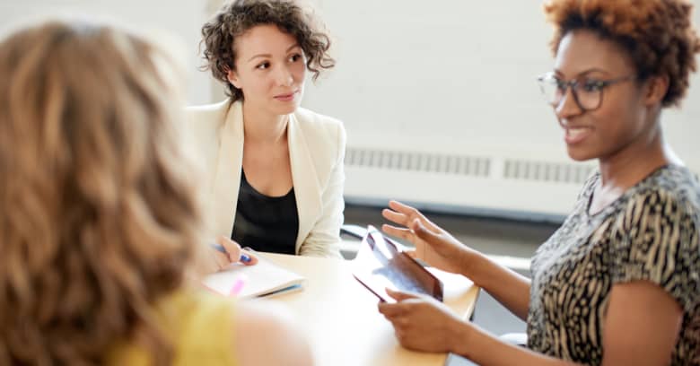 3 women sit at a table, one holds a tablet. They discuss how they are going to invest their money.