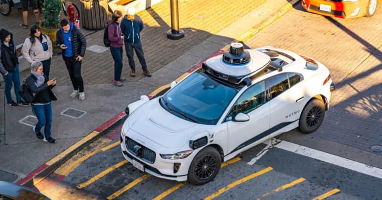 A crowd of people gather to look and take pictures of a self-driving car that is parked on a public street.