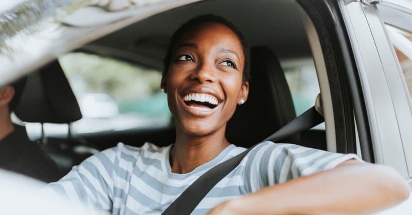 A cheerful woman in a striped shirt is behind the wheel of a car, smiling broadly.