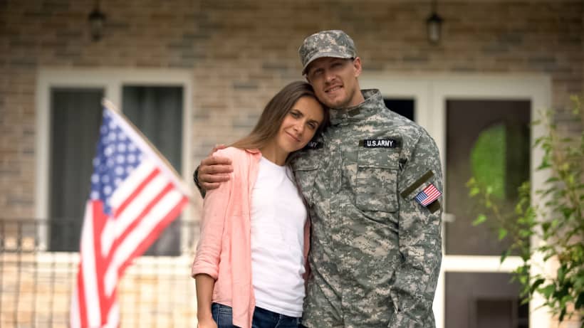 U.S. Army soldier in uniform stands with his arm around a woman in front of a house.