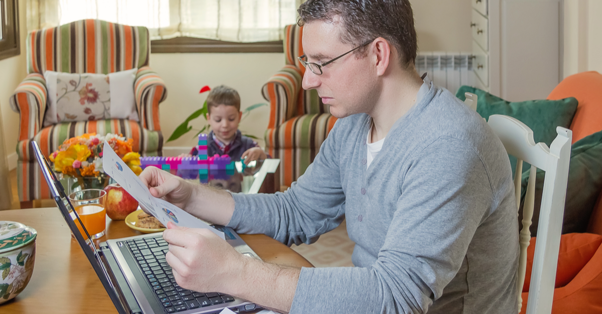 a man sits at a table while working on his computer while his son plays with colorful blocks in the background.