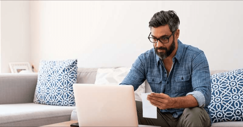 A man sits on his couch and looks at his computer and his medical bills.