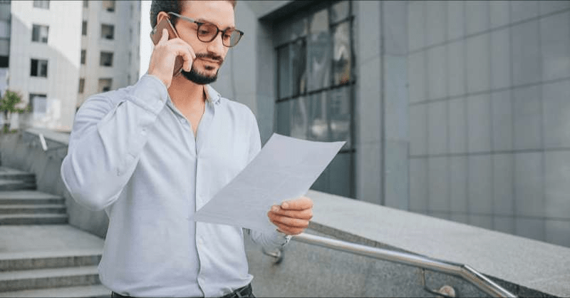 A man speaks on the phone while looking at a medical bill. He is standing outside on some concrete steps next to a building.