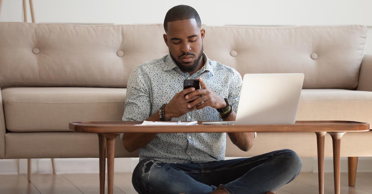 A man sits on the floor in front of his coffee table looking at his phone as he takes a break from freelance work.