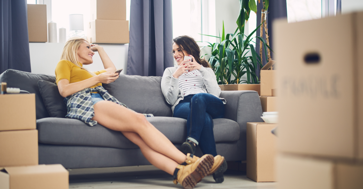 Two young woman, surrounded by moving boxes, sit on the couch in their new apartment