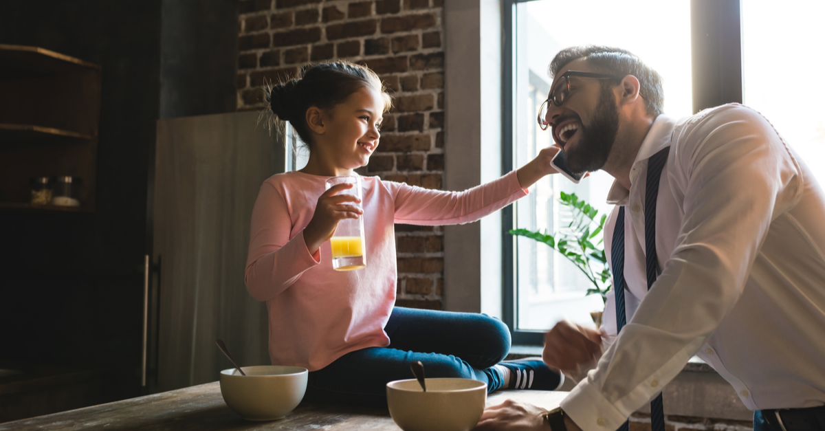 A man and his young daughter share a phone call together in the kitchen during breakfast