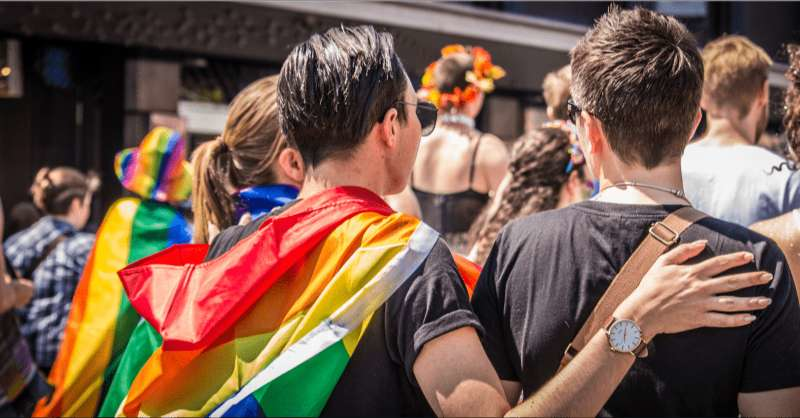 Two people are seen from behind with a rainbow flag as the watch a pride parade