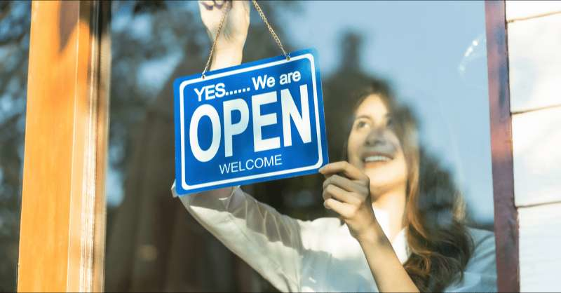 A woman hangs an 'open for business' sign in a window.