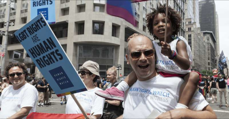 an LGBTQ+ dad marches in a pride parade with his daughter sitting on his shoulders